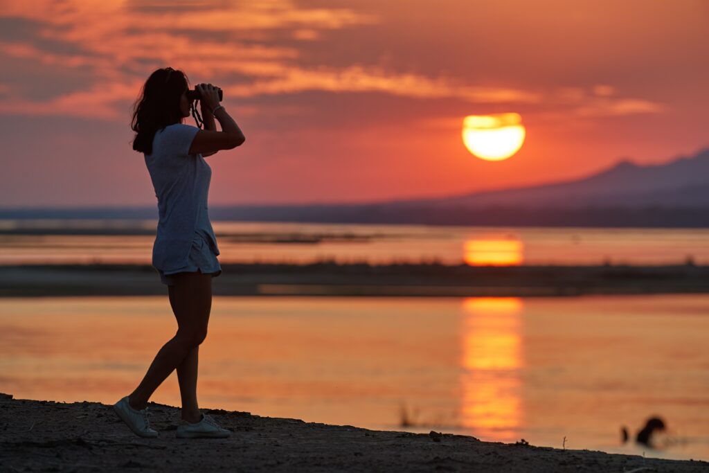 Silhouet van een vrouw met verrekijker aan de oever van de Zambezi in Zimbabwe tijdens zonsondergang – safari met Christoffel Travel